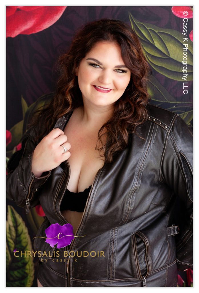 heterochromia iridis Curly brunette is a Beauty Beyond Size standing against flower wall in leather jacket for Boudoir Photoshoot