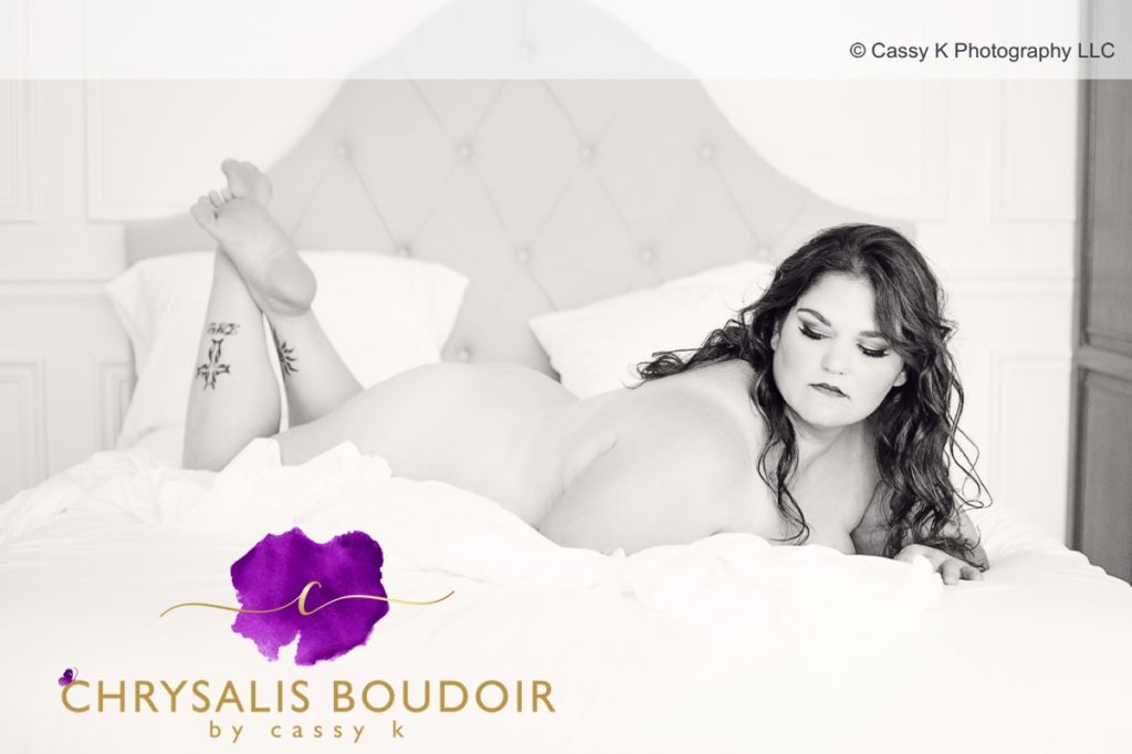 Curly brunette is a Beauty Beyond Size laying on stomach with a white sheet only for Boudoir Photoshoot