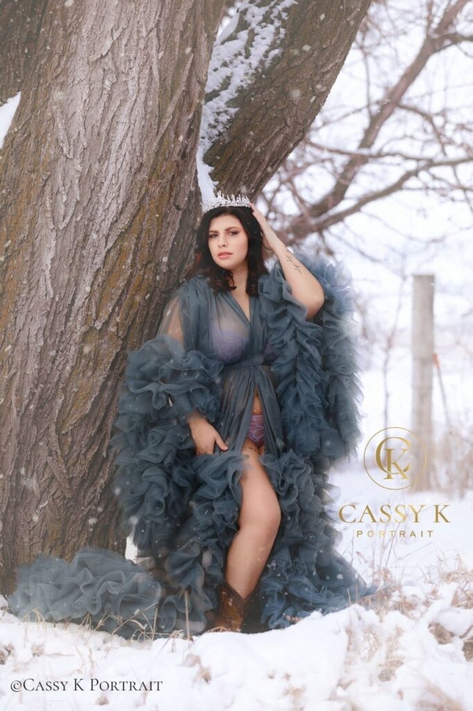 Young woman channelling ice princess vibes works through body changes and falls in love with herself wearing ice crown and gray tulle couture gown in the snow during boudoir photo shoot