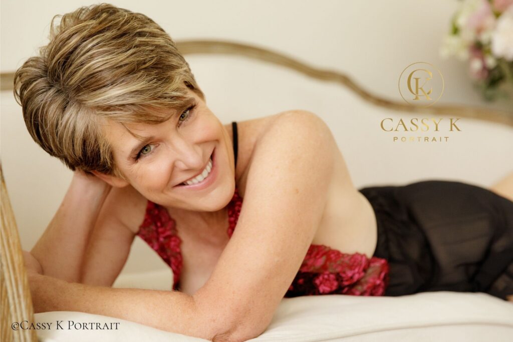 A radiant woman with short blonde hair smiling while resting on white bedding in red lace lingerie, beautifully showcasing how to embrace sexy at any age.