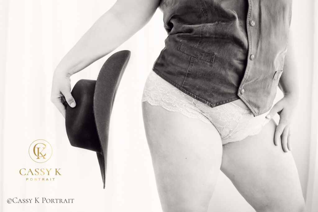 Black-and-white close-up of a woman wearing a leather vest and lace underwear while holding a cowboy hat.