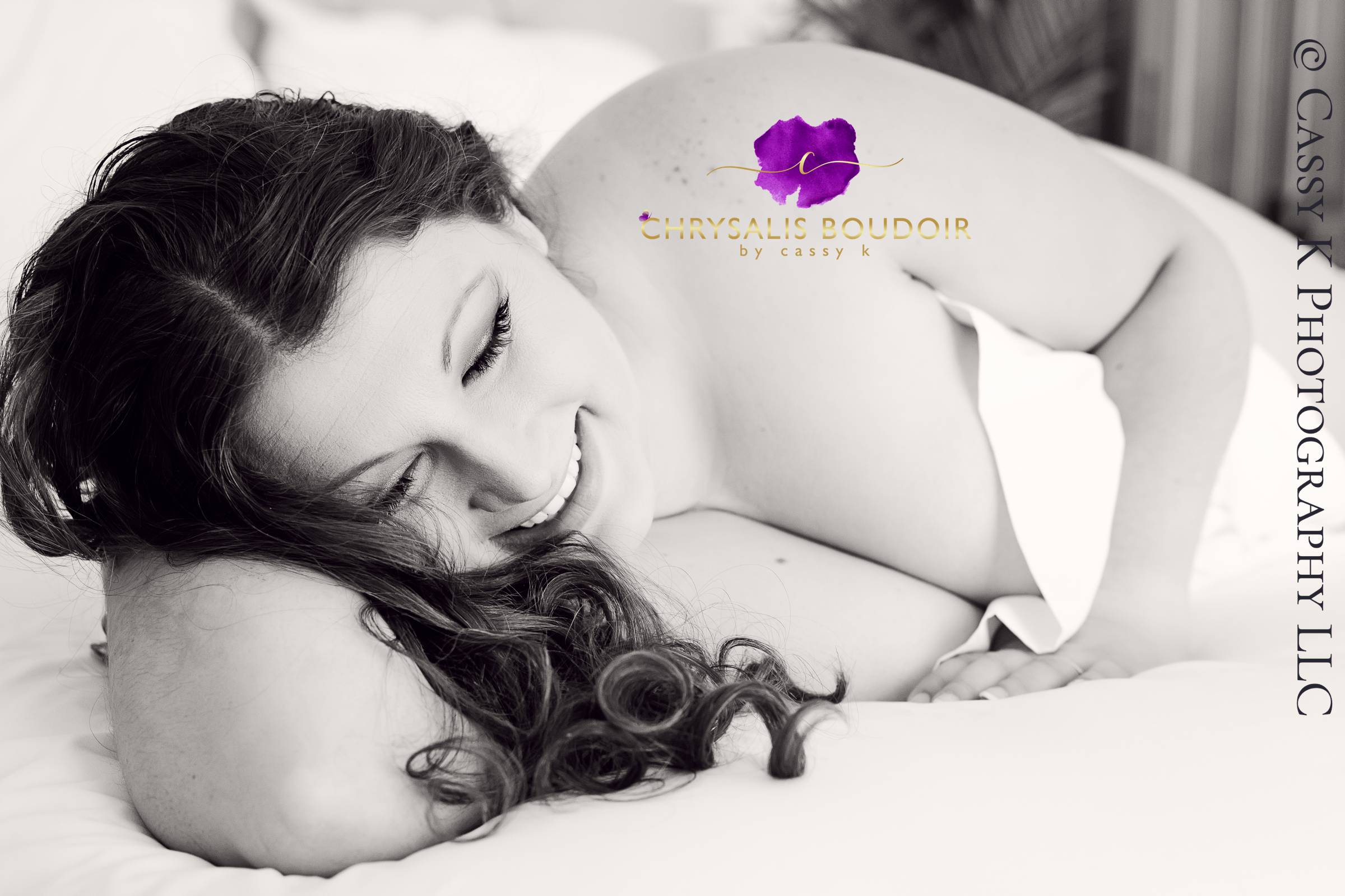 Curly Brunette, brown eyed woman giggling on the bed during white sheet Boudoir Photoshoot to celebrate her mom bod
