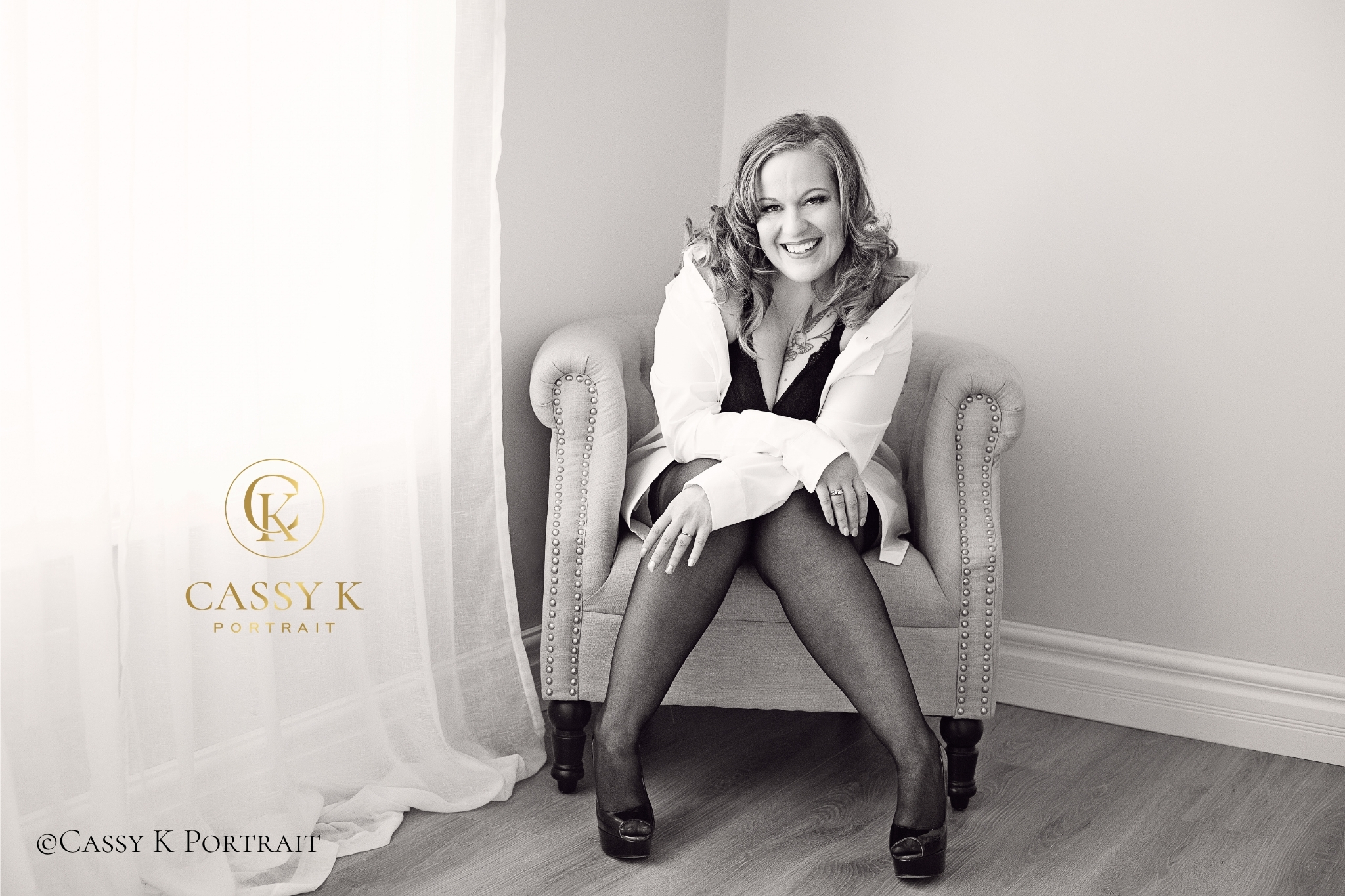 Smiling woman seated in an armchair wearing a white button down shirt, black outfit, and heels during her studio portrait session.