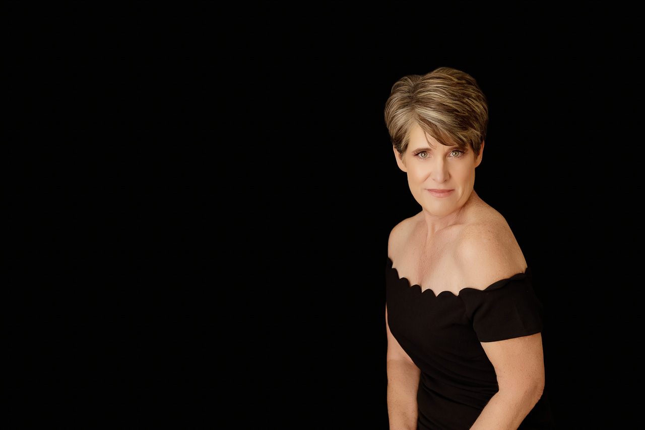 A stunning portrait of a woman with short hair wearing a black scalloped off-the-shoulder dress against a dark studio background during an over 40 photoshoot.