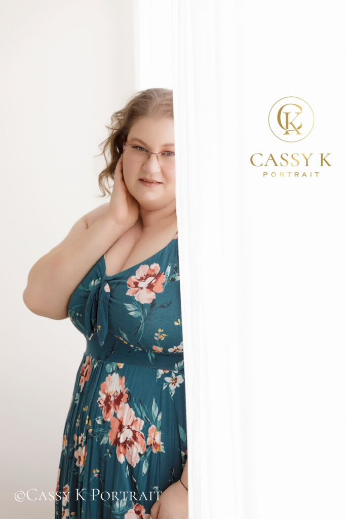 Woman in a teal floral dress posing with one hand at her neck beside a white curtain.